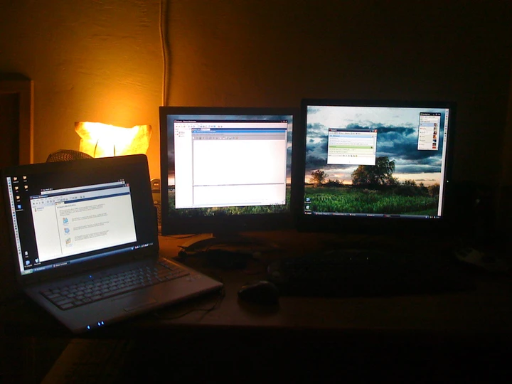 A laptop and two monitors glowing in a dark home workstation